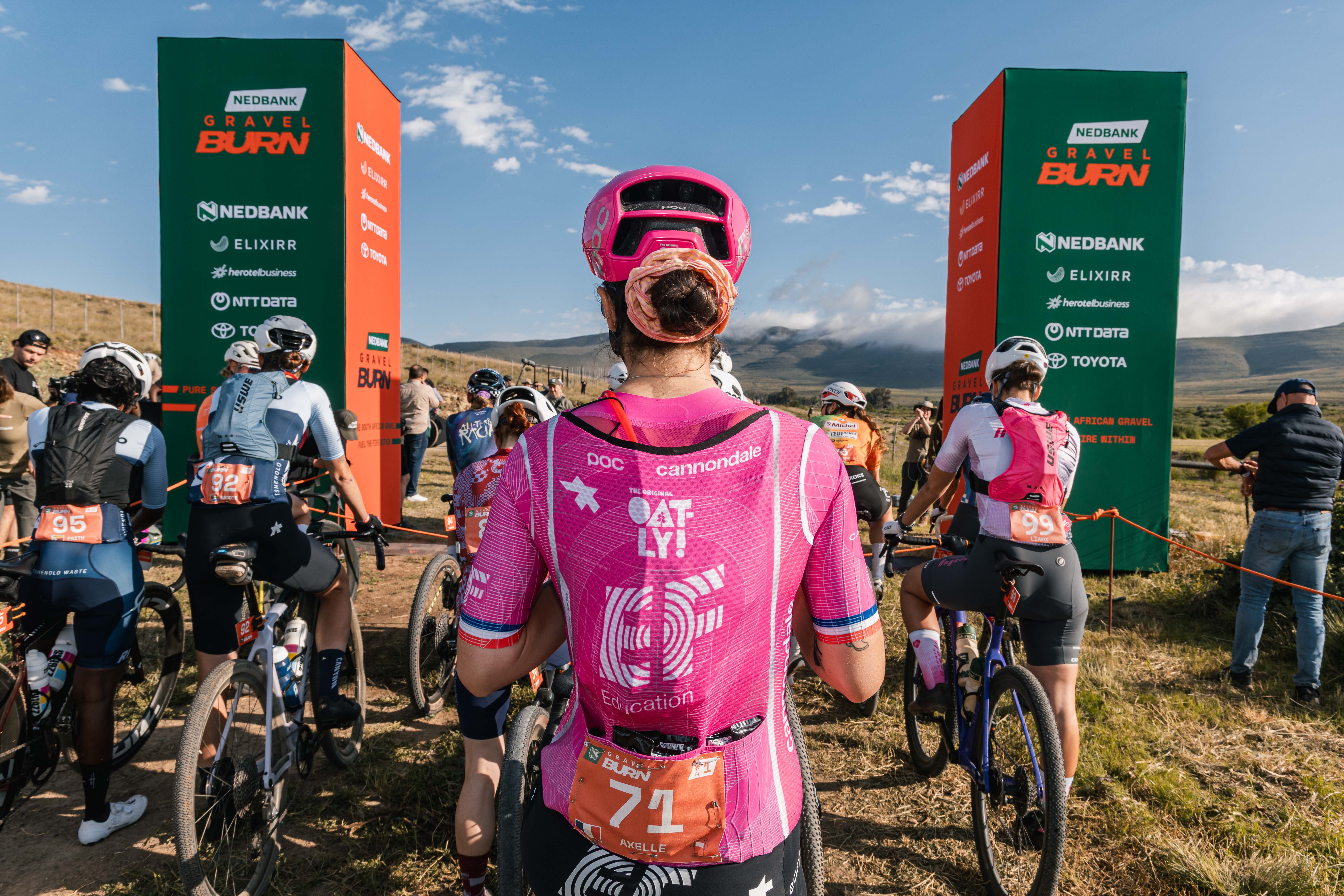 Cyclists line up at a Nedbank Gravel Burn start arch, with a rider in a pink Canyon jersey seen from behind in the foreground.