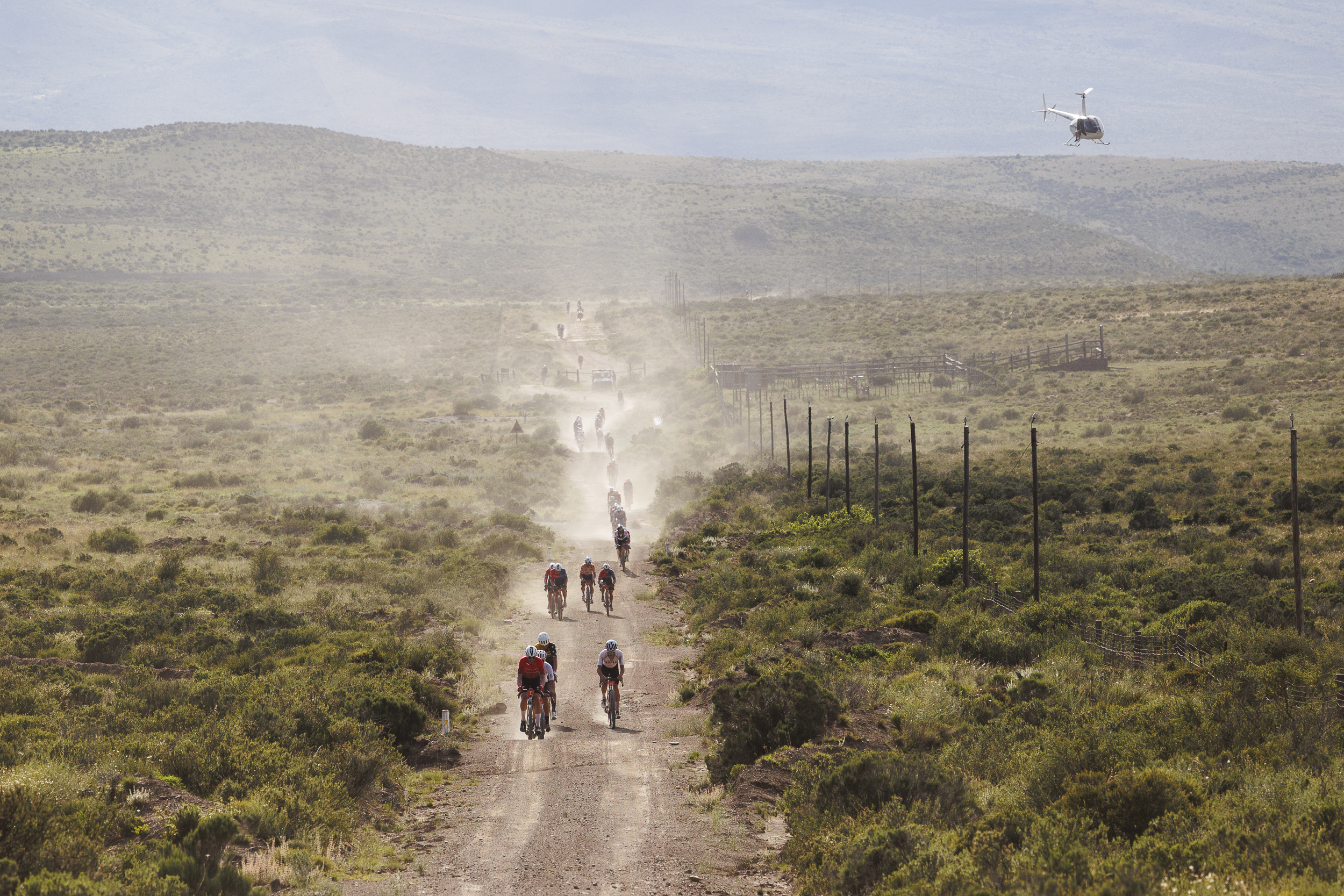 Cyclists ride along a dusty gravel road through open scrubland, with a helicopter flying above and hills in the background.