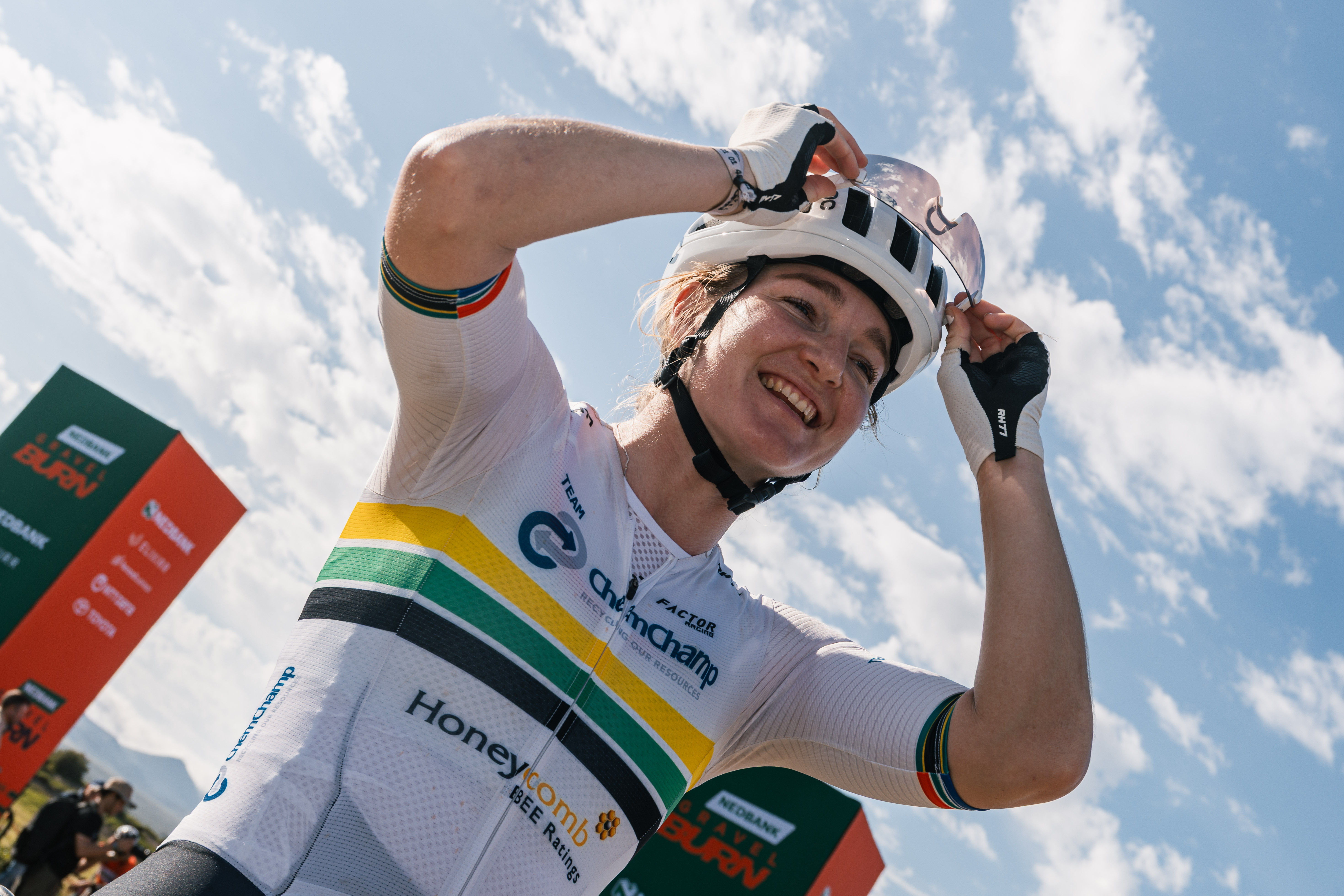 A smiling cyclist in a white national champion-style jersey adjusts a helmet near Nedbank Gravel Burn event signage under a blue sky.