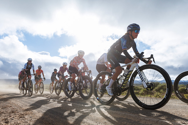 A group of gravel cyclists ride along a dusty road under a bright cloudy sky.
