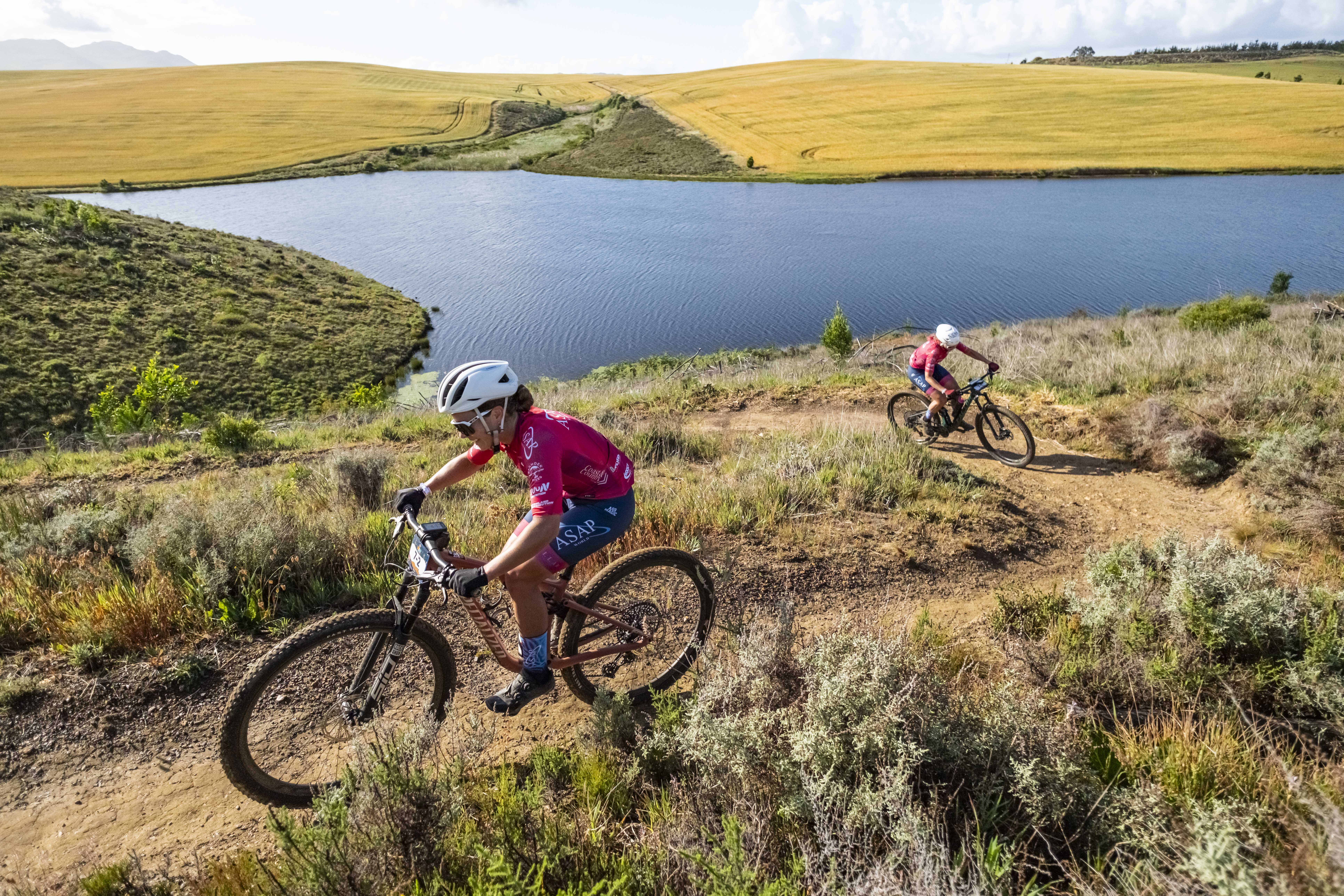 Two mountain bikers ride along a dirt trail above a dam in open countryside.