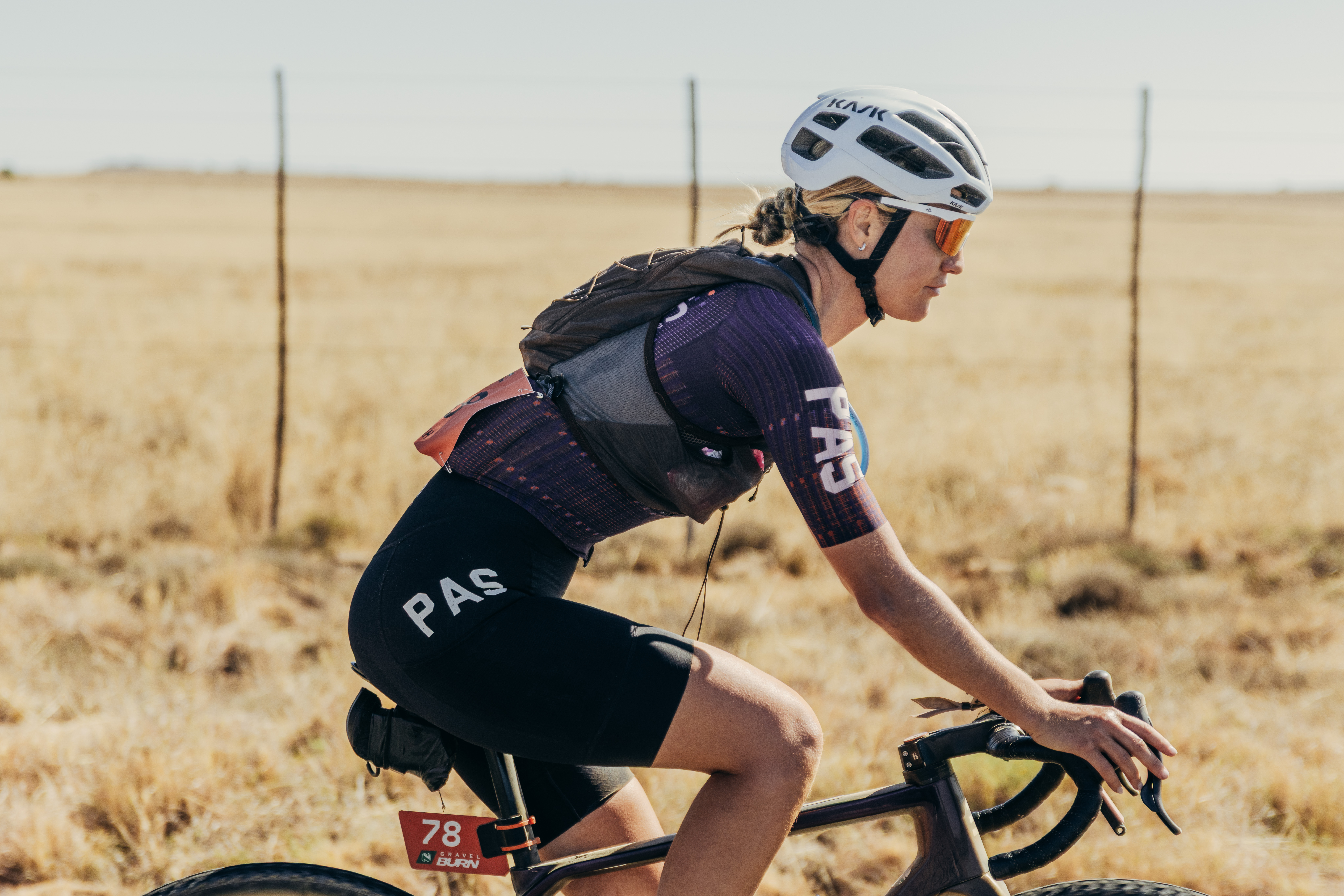 A cyclist in a helmet rides a gravel bike past dry grassland and a fence.
