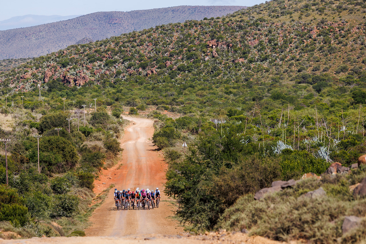 A group of cyclists rides along a dusty gravel road through a hilly semi-desert landscape.