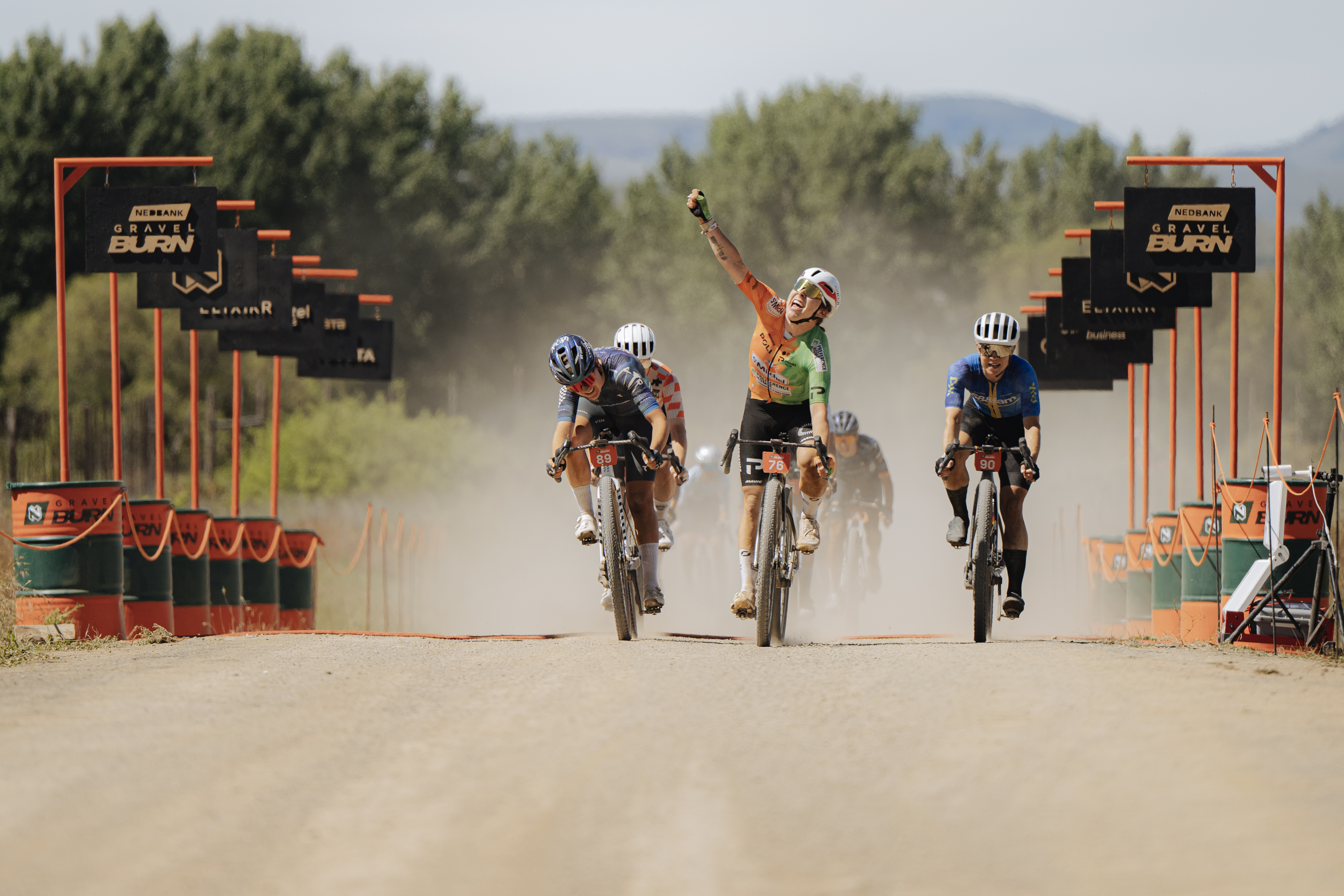 Three cyclists sprint on a dusty gravel road under Nedbank Gravel Burn finish gantries, with the centre rider raising one arm.